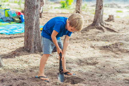 Camping people outdoor lifestyle tourists, summer forest near lazur sea. Blond boy play study survival techniques, practice methods to dig the earth with a shovel. Natural children educationの写真素材