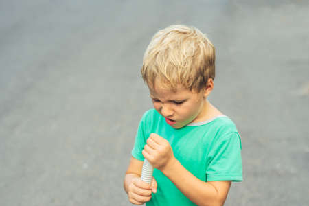 Funny mischievous cute blond boy with freckles face squints, looks into plastic corrugated pipe hose, artistic emotions. Behaviour education problems, simple joys of happy childhood, people conceptの写真素材