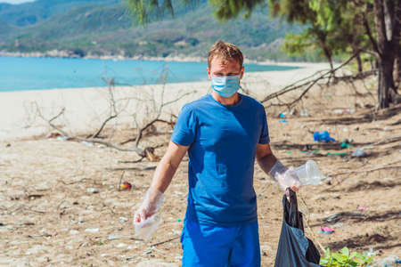 Volunteer man in face mask pick up garbage pollute beach near sea, hold look at discarded disposable plastic cups. Problem of spilled rubbish trash, planet pollution environmental protection conceptの写真素材