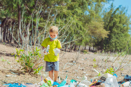 Volunteer boy in face mask helps to pick up garbage which pollute beach near forest. Problem of spilled rubbish trash planet pollution environmental protection concept. Natural children educationの写真素材