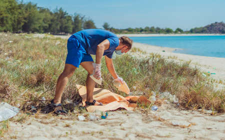 Volunteer in blue face mask lazur sea sand beach. Active man pick up garbage plastic foam bags film. Spilled rubbish trash planet pollution environmental protection. Natural social problems conceptの写真素材