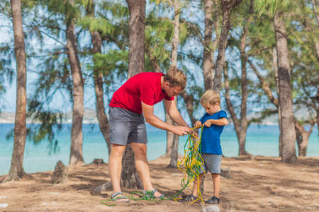 Camping people outdoor lifestyle tourists in summer forest near lazur sea. Blond boy son with father study survival techniques, practice methods of tying rope knots. Natural children educationの写真素材