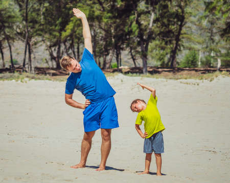 Sport for little children. Handsome father and cute son exercising on sand beach near forest park, having workout enjoying together in the fresh air, correct squatting and side bending techniqueの写真素材