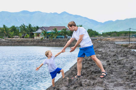 Dad helps son go down to lake water, explains safety rules. Family together walk play. Home natural child education, fathers day, fatherly responsibilities, influence on formation of son worldviewの写真素材
