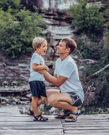 Father hugs tickle son sitting squatting, smile. Happy childhood, fathers day, mountain, chain background as symbol of strong bond between dad and child. Father influence on son worldviewの写真素材