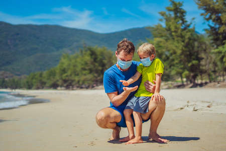 Father squatting holding son looking on open hand, play compare their palms, wearing face blue mask protect coronavirus walking nature sand sea beach near forest park sun day. COVID normal realityの写真素材