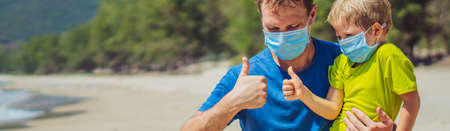 Banner Father squatting holding son playing fists with thumbs up, wearing face blue mask protect coronavirus walking nature sand sea beach near forest park sun day. COVID Global pandemic virus realityの写真素材