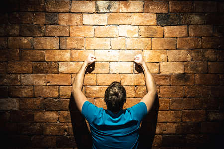Rear view man under life crisis problems stand facing leaning against brown brick wall pounding smashing , hands raised up, wants to overcome obstacle, reach a goal, leader, motivation. Looking upの写真素材