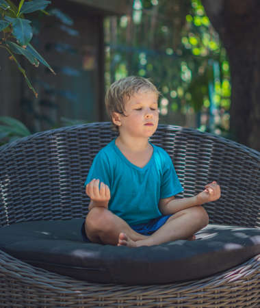Calm little male boy sit on couch outside garden practice yoga. Young generation modern child rest relieve negative emotions with closed eyes in lotus pose at sunrise. Mindfulness meditation conceptの写真素材