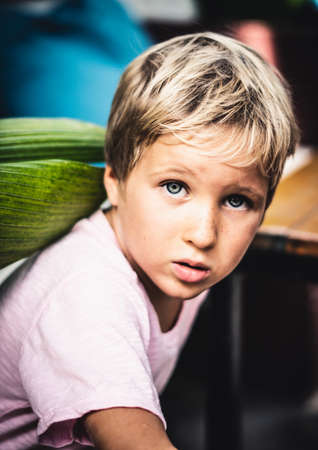 Portrait cute freckled blond blue eyed boy serious mood emotion facial expression look at camera think in pink, hide under leaves. Childhood problems relationship, harassment physical abuse conceptの写真素材