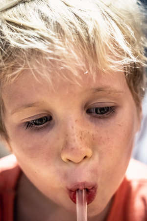 Close up portrait blond freckled thirsty child boy serious concentrated facial expressions drinking from straw, looking eyelashes down, orange t-shirt. Childhood real people conceptの写真素材