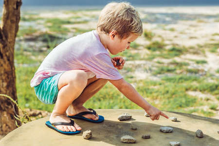 Cute Child freckled face boy play squatting outside nature sun day, driving traffic regulation rules use stone imitation cars toy. Expressive Serious face point Finger gesture. Education imaginationの写真素材