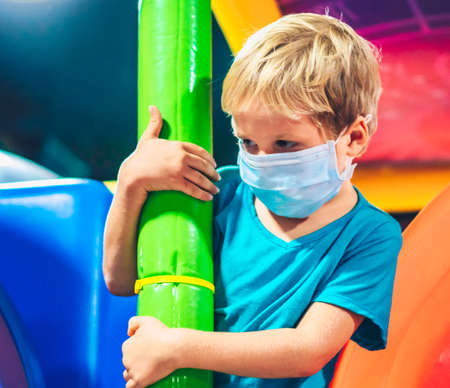 Cute blond child boy, amusement playground entertainment indoor center, wraps arms around hold hands, look far away sad wear blue protective mask new lifestyle due to COVID 19 Coronavirus wait changeの写真素材