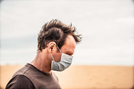 Side portrait young man wear protective mask, face expression emotion look down dune. Concept COVID stress syndrome, mental health, fear of danger contamination, adverse socio economic consequenceの写真素材