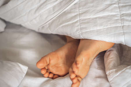 Close-up side view child boy two feet stick out under white blanket on bed. Sleep and relax concept,...