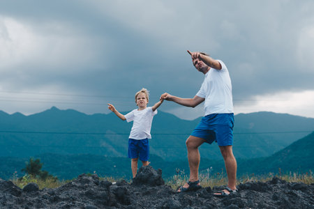 Father son or adult younger brother walk. Cloudy sky before rain, mountain background. Man show points finger into distance, serious boy look far away. Solve problems together. Natural educationの写真素材
