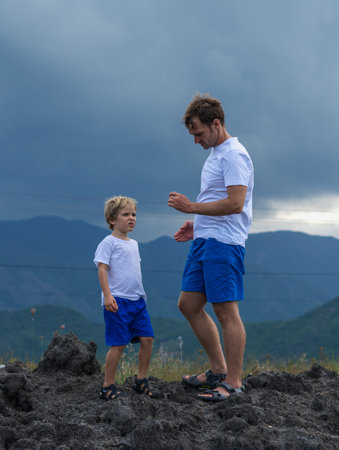 VERTICAL Serious Father son or adult younger brother walk together. Cloudy sky before rain, mountain background. Man found holds unusual stone in hand, boy attentive look. Natural science educationの写真素材