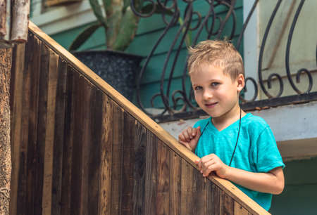 Lifestyle portrait real skin facial expression caucasian little boy stand outside hold railing stairs. Wood board Copy space. Play mischievous intriguing look. Happy childhood games kid productsの写真素材