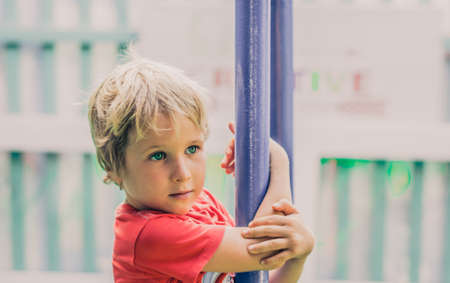 Close up lifestyle side photo. Portrait little boy happy look far away right distance hanging on pipe sport metal horizontal bar hands clasp keep. Outside games children Protection Day, playgroundの写真素材
