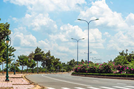 Asphalt curve empty highway, flower bushes as divider, white lines marks, lighting poles lanterns, green trees on sidewalk, blue sky cumulus clouds sun day. Beautiful summer landscape. Travel tripの写真素材