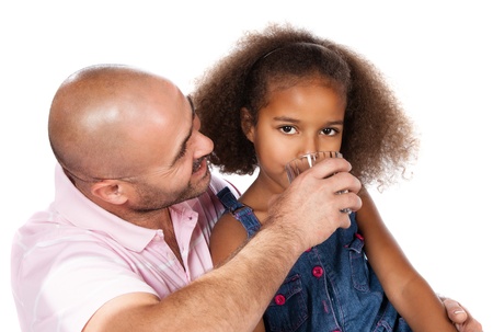 Adorable cute african child with afro hair wearing a denim dress. Her father is helping her to drink water from a clear glass.の写真素材