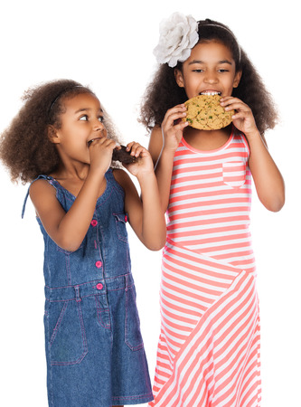 Two adorable cute african children with afro hair. One is wearing a white and pink striped dress and the other a denim dress. Each has a big choc chip cookie.の写真素材