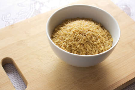 White bowl of uncooked brown rice on a wooden chopping board.の写真素材