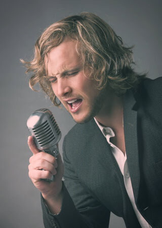 Portrait of a handsome caucasian male singer wearing a white buttoned shirt and a grey jacket. The man is singing into a silver performance microphone.の写真素材