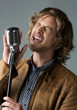 Portrait of a handsome caucasian male singer wearing a blue checkered button shirt and a leather jacket. The man is singing into a silver performance microphone.の写真素材