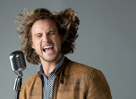 Portrait of a handsome caucasian male singer wearing a blue checkered button shirt and a leather jacket. The man is singing into a silver performance microphone.の写真素材