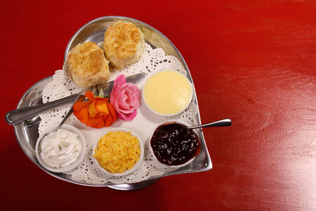 Delicious butter scones on a silver heart shaped tray served with roses cheese cream and jam preserves on a red wooden table.の写真素材