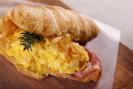 Delicious croissant with bacon scrambled egg caramelised onions and rosemary served on a wooden plate on a wooden table.の写真素材