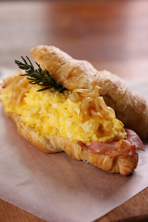 Delicious croissant with bacon scrambled egg caramelised onions and rosemary served on a wooden plate on a wooden table.の写真素材