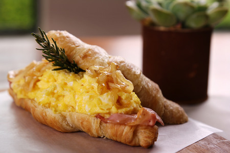 Delicious croissant with bacon scrambled egg caramelised onions and rosemary served on a wooden plate on a wooden table.の写真素材