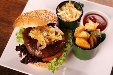 Delicious juicy beef burger with caramelised onions and salad served with potato wedges on a white plate on a wooden table.の写真素材