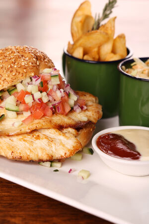 Delicious chicken burger with chopped salad served with potato wedges on a white plate on a wooden table.の写真素材