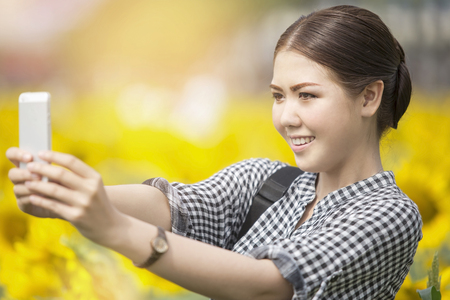 Young happy Asian woman smiling while taking selfie picture with mobile phone in sunflowers fieldの写真素材