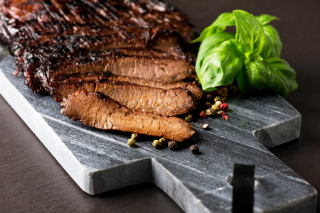 Close up sliced medium grilled beef steak with pepper and oregano leafs on marble meat cutting board on dark stone background