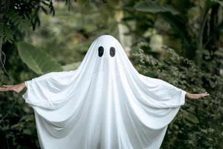 A young boy dressed as a ghost for Halloween wears a white sheet with two black eye holes. He raises both arms in a spooky gesture, surrounded by dark green foliage