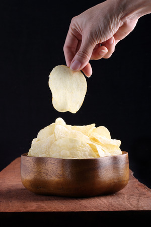 Crispy potato chips in wooden bowl with hand picked on wooden tray and black background.の写真素材