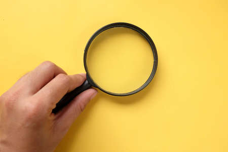 top view and flat lay man hand holding black a magnifying glass on the yellow office desk backgroundの写真素材