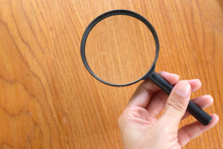 copy space woman hand holding black a magnifying glass on the brown wooden office desk backgroundの写真素材