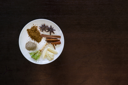 various herbs cinnamon, star anise, sarlic, black pepers, corriander root  in white plate on black brown wooden backgroundの写真素材
