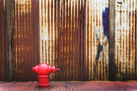 red fire hydrant on footpath over rusty corrugated iron metal fence zinc wall background with copy spaceの写真素材