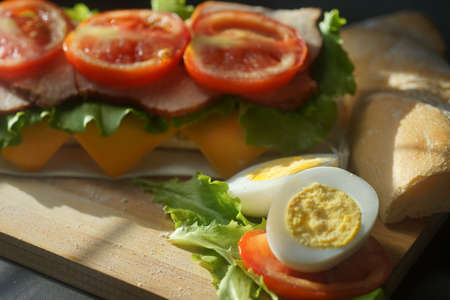An appetizing sandwich with ham, cheese, tomatoes and lettuce lies on a board with egg and lemon on black backgroundの写真素材