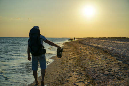 a guy with a backpack walks along the coastline in the evening barefoot on the sand, holds sandals...