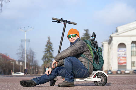 Guy photographer sits on an electric scooter in the spring in the park. He is wearing a warm jacket and hat. Nearby lies snow and coldの写真素材