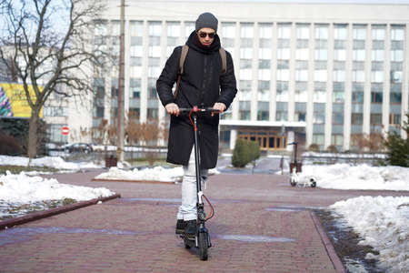 The guy rides an electric scooter in the spring in the park. He is wearing a warm jacket and hat. Nearby lies snow and coldの写真素材
