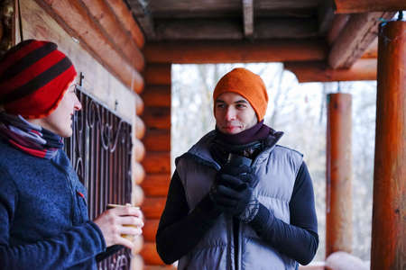 Two guys chat after skiing. They stand on the balcony of a wooden house in winter clothes, drink tea...