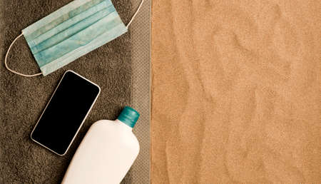 Summer concept on the beach, gray towel with virus protection mask, black screen of the smartphone,...
